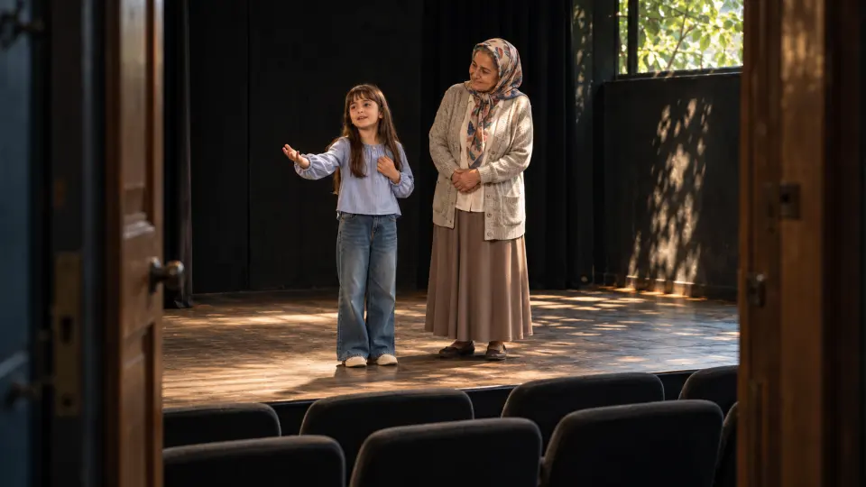 Kapıdan görünen tiyatro sahnesinde, başörtülü büyükannesinin yanında oyunculuk alıştırması yapan küçük kız.