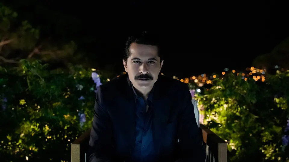 İsmail Hacıoğlu, gece ışıkları önünde takım elbiseyle oturuyor (NOW röportaj görseli).