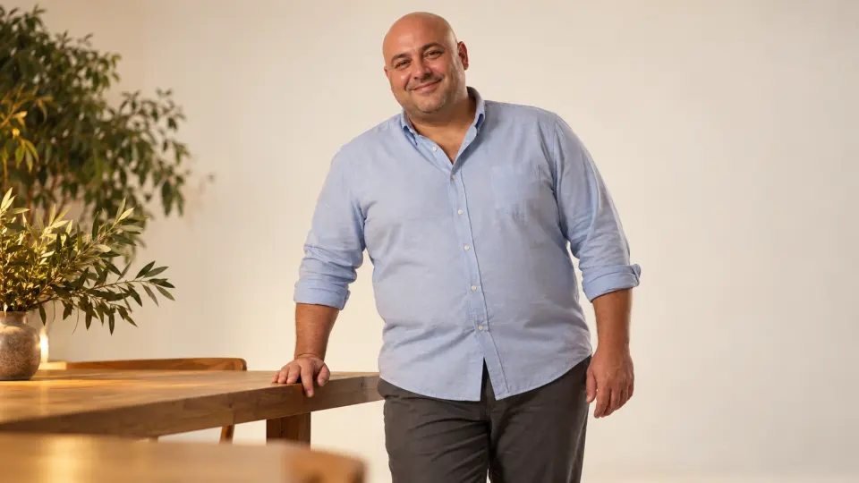 Beyaz stüdyoda mavi gömlekli, tıraşlı başlı büyük beden erkek model, ahşap masa yanında gülümseyerek poz veriyor.